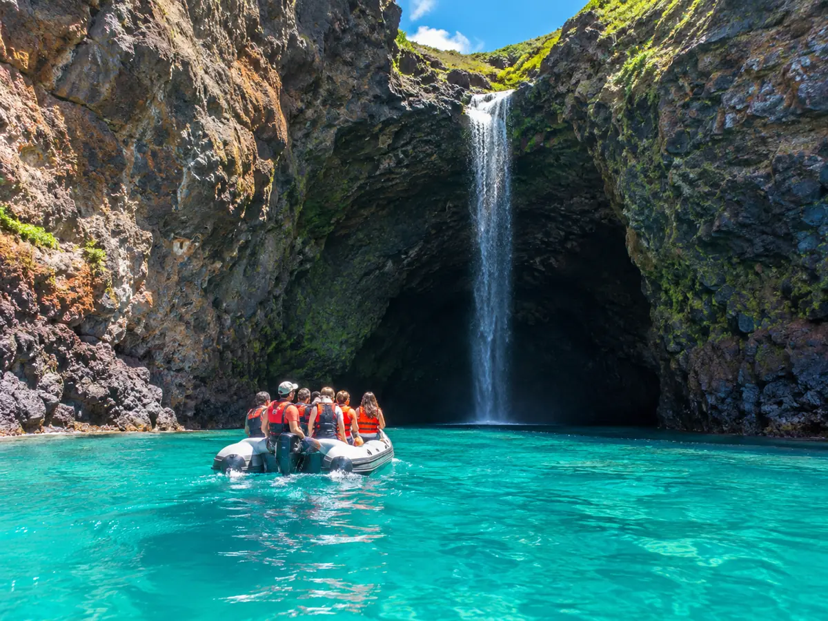 Ein Schlauchboot gleitet unter einem schmalen Wasserfall in eine hohe Lava-Meereshöhle